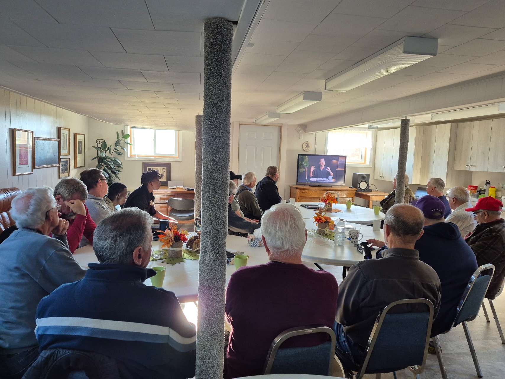 Men gathered around tables in the church basement, watching a teaching video on the TV